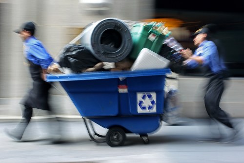 A blue waste collection bin on wheels, positioned on a paved street or driveway, is filled with assorted rubbish including a black plastic bag, a large green garden waste bin, and various other debris such as cardboard boxes and plastic containers. The bin is in motion, with two workers wearing navy and light blue uniforms on either side, pushing it forward. The background is blurred, indicating movement, and the scene occurs in an urban area, possibly within the vicinity of Hendon, North London, as suggested by context. This image relates to rubbish removal services offered by Garden Clearance Hendon, illustrating a busy and efficient collection process typical of professional waste clearance operations.