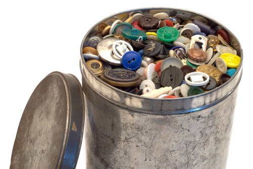 A close-up image of a cylindrical metal rubbish bin filled with a variety of assorted buttons, including plastic, fabric-covered, and metal types, in different sizes and colors such as black, white, blue, green, yellow, brown, and beige. The bin is positioned outdoors on a concrete surface with a slightly rusted and weathered finish, and its matching metal lid is leaning against the side of the bin. The background is plain and white, keeping the focus on the densely packed collection of buttons inside the bin, which suggests the contents are related to textile or craft waste, relevant to rubbish removal services in Hendon or similar areas in London.