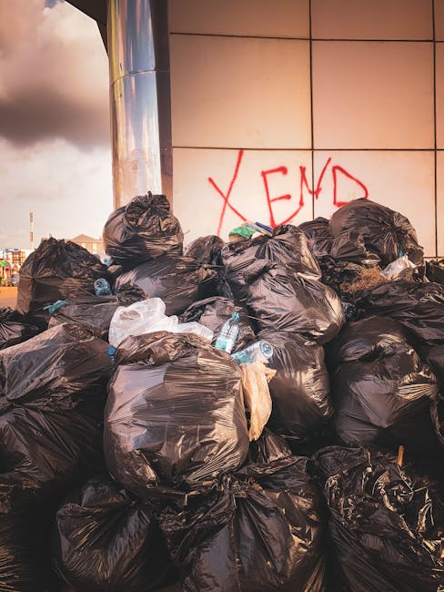 A pile of mixed rubbish including black plastic bags, a yellow plastic tray, and an old, dirty car tire, all casually strewn on a gravel surface in an outdoor setting. The rubbish is positioned in fro