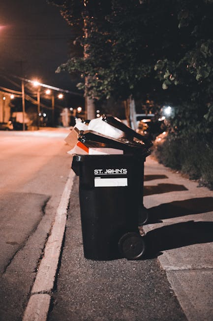 Three large black plastic rubbish bags, filled and tied at the top, are resting on a paved sidewalk next to a black metal fence. The bags appear to contain household waste or general rubbish, characte