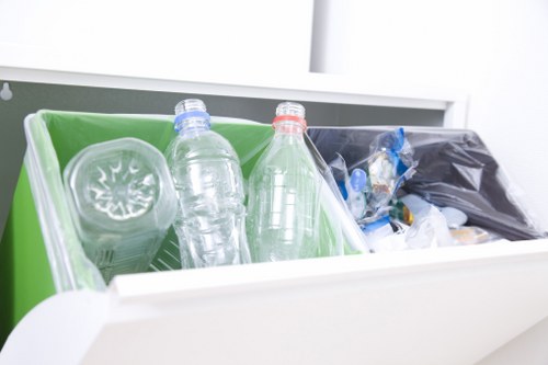 A close-up view of an open white drawer containing several discarded plastic bottles and packaging materials, including a transparent plastic bottle with a blue cap, another with a red cap, and various smaller plastic wrappers and containers. The interior of the drawer features a green and black compartmentalized storage box, used for organizing rubbish and recyclables. The background shows a clean, modern surface and minimal surroundings, indicative of a household or office environment in Hendon, suitable for promoting rubbish collection and waste disposal services by Garden Clearance Hendon. The focus is on the clearly visible plastic waste items positioned to highlight typical domestic rubbish ready for disposal or collection, reflecting the service category of rubbish removal in the local area.