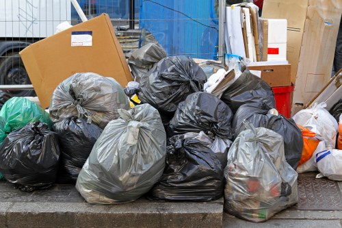 A collection of tightly packed black and grey rubbish bags filled with waste material, stacked along the edge of a pavement or driveway, with some bags leaning against a metal curb. The bags are made of plastic, with some appearing slightly crinkled or glossy, and others more matte in finish. In the background, there are cardboard boxes and various yard or household waste items, suggesting clearance activity, possibly arranged for collection by a professional rubbish removal service such as Garden Clearance Hendon. The scene is outdoors, likely in an urban residential area near Hendon, with a concrete surface and some construction or garden debris visible. The overall environment indicates a typical rubbish clearance process, with the waste awaiting disposal or collection, reflecting the company's service of managing garden or household rubbish removal efficiently in the local area.